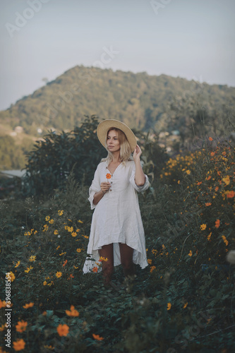 Obraz na płótnie The blonde in a linen dress and straw hat walks among herbs and wildflowers on a meadow in the mountains. A woman in the Boho style, resting in the countryside, a simple village lifestyle w salonie