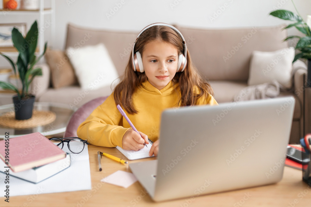 Homeschooling. Smiling girl learning from home while covid-19 pandemic, doing homework, using headphones and laptop for distance learning. Remote school, online education during coronavirus quarantine Homeschooling. Smiling girl learning from home while covid-19 pandemic, doing homework, using headphones and laptop for distance learning. Remote school, online education during coronavirus quarantine