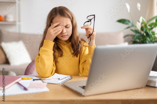 Tired school girl rubbing her eyes, holding eyeglasses, exhausted from studying at home and doing homework, sitting in front of laptop with notebook. Homeschooling, distance education due to covid-19