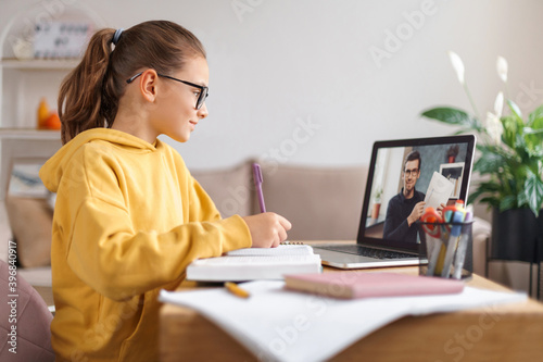 School girl having video conference with online teacher on laptop in living room at home. Homeschooling, distance learning, online education due to coronavirus pandemic. Side view. E-learning concept photo