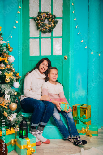 Mom and daughter are sitting on the steps near the Christmas door and waiting for the new year. A loving family, they cut the Christmas tree and give gifts. © Вероника Преображенс