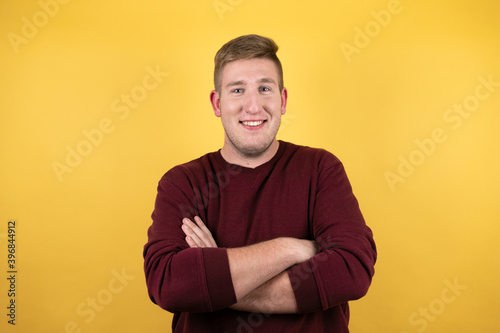 Young blonde man wearing a casual red sweater over yellow background smiling confident showing teeth with arms crossed