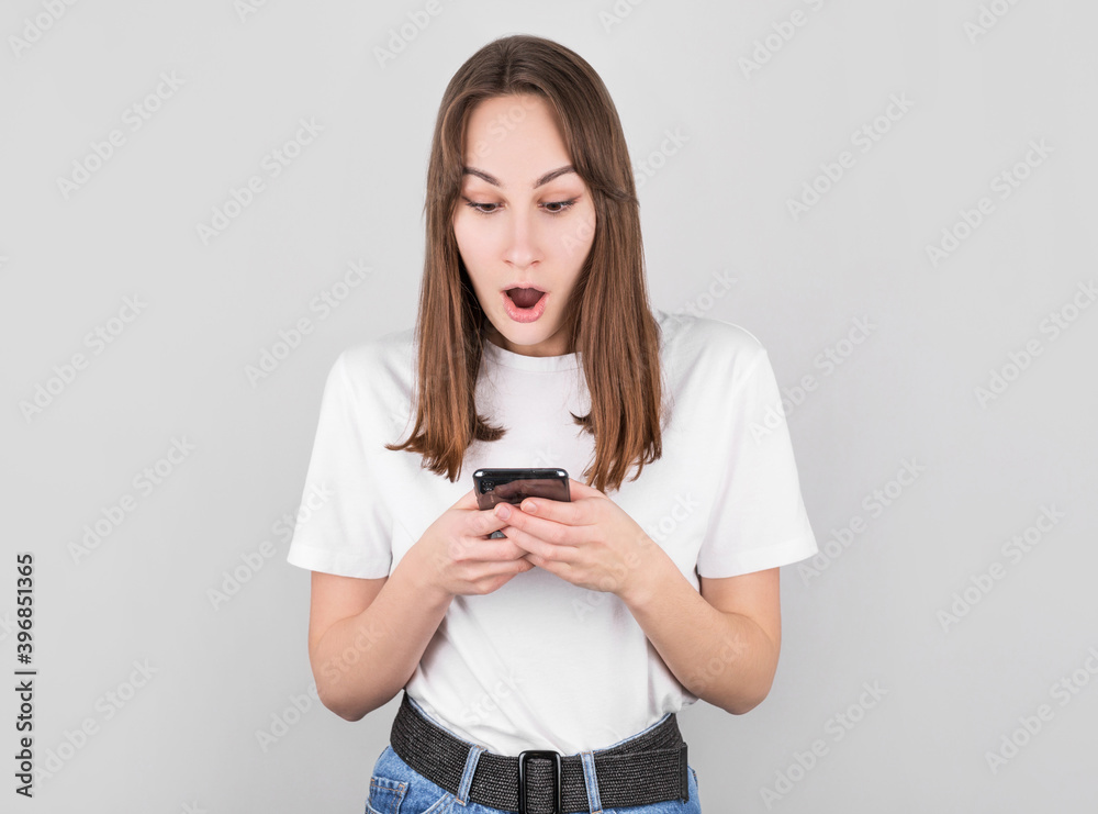 Portrait if a shocked young girl looking at mobile phone isolated over gray background Portrait if a shocked young girl looking at mobile phone isolated over gray background