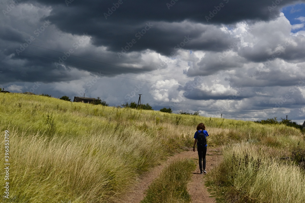 Anonymous young girl walking on the dirt road through Ukrainian steppe with a stunning cloudscape. Windy weather with overcast sky in springtime. Traditional rural landscape with  in Ukraine