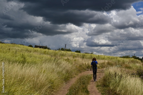 Anonymous young girl walking on the dirt road through Ukrainian steppe with a stunning cloudscape. Windy weather with overcast sky in springtime. Traditional rural landscape with in Ukraine