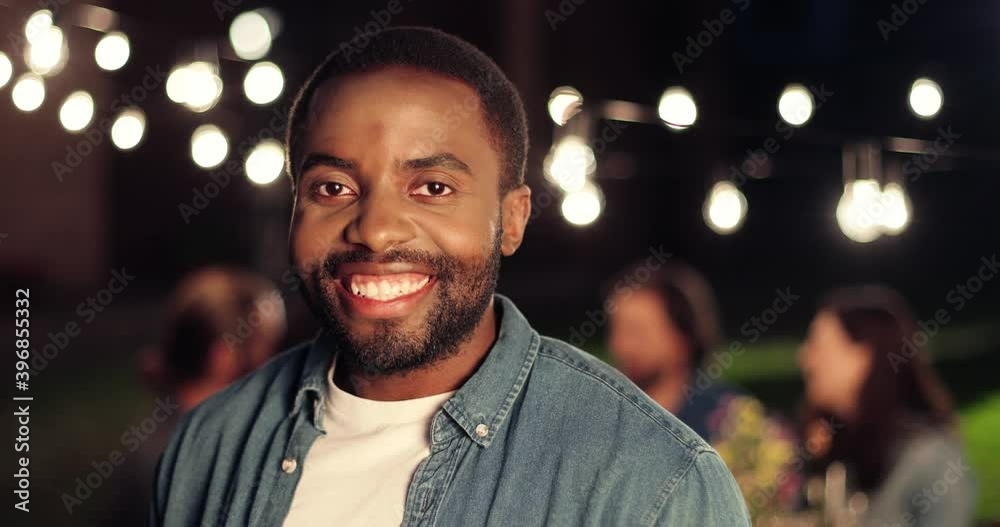 Portrait of young handsome African American man smiling cheerfully and standing at outside party at night. Happy guy at evening celebration. People at table on background.