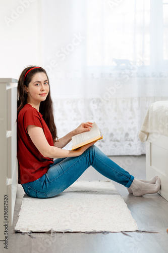 A young Caucasian woman is sitting on the floor and leafing through a book. Copy space. Side view. In the background-the white interior of the room. Concept of education and world book day