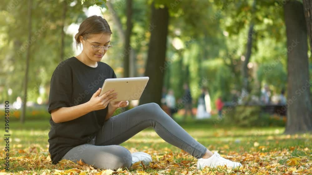 Slow Motion Young girl in glasses sits on the lawn in autumn city park and looks photos on his tablet computer