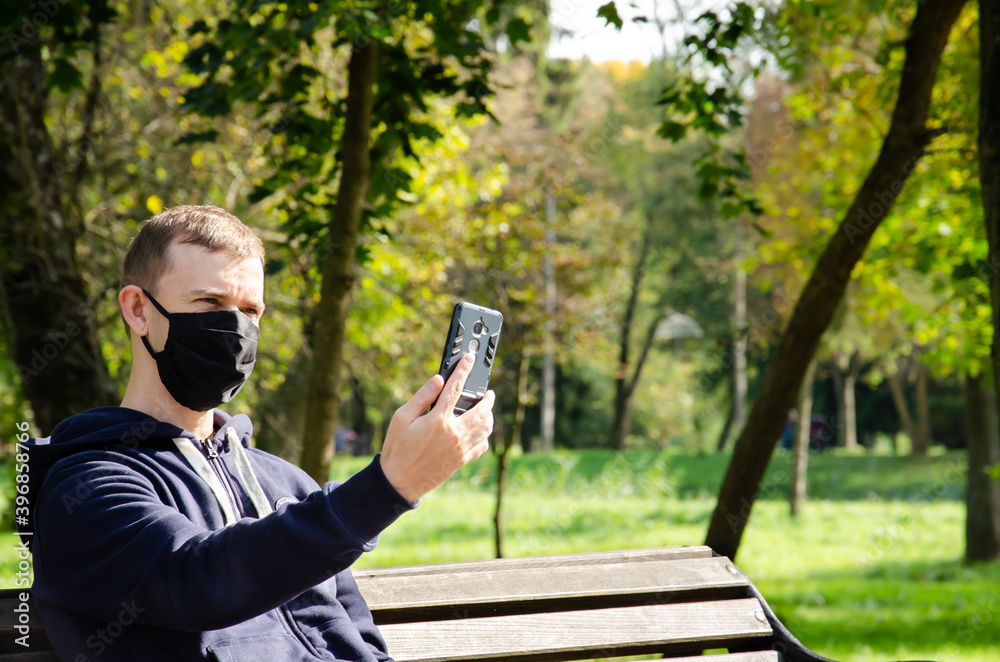 Fototapeta premium Caucasian man in protective medical mask making video call on phone and sitting on bench in the park during coronavirus outbreak with copy space. New reality. Long distance relationship concept.