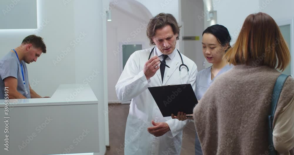 Portrait of Caucasian male doctor standing in hospital and speaking with Asian female nurse about treatment results looking at medical history. Healthcare professionals talking in clinic Medic concept Portrait of Caucasian male doctor standing in hospital and speaking with Asian female nurse about treatment results looking at medical history. Healthcare professionals talking in clinic Medic concept