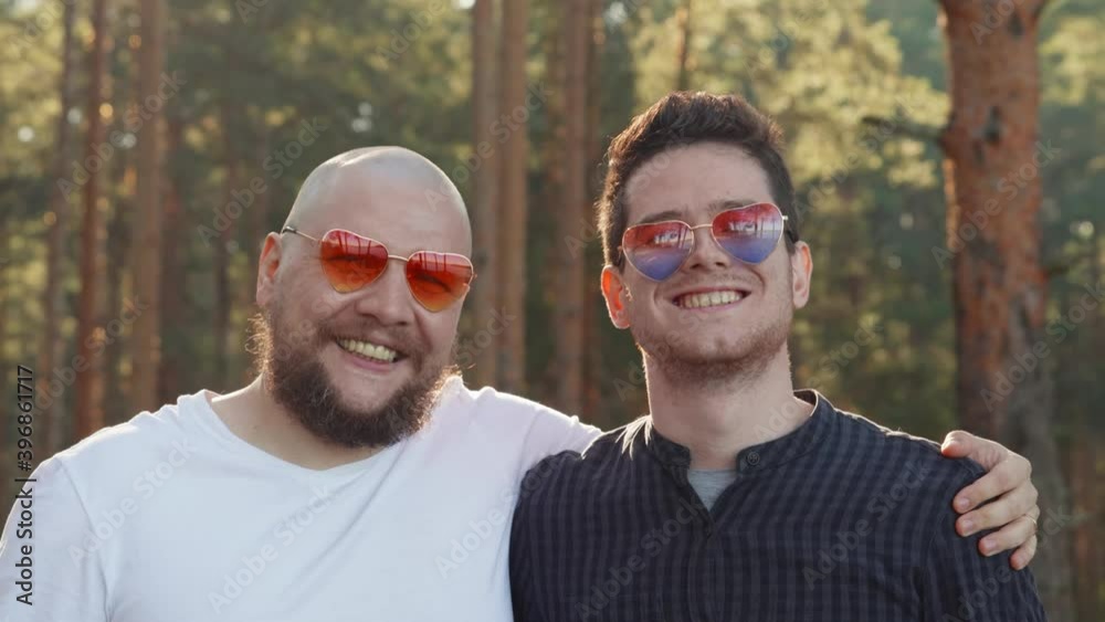 Portrait of two affectionate gay men in love wearing sunglasses of heart shape smiling to camera. Homosexual couple dating in nature. LGBTQI, Pride Event, LGBT Pride Month, Gay Pride Symbol. Portrait of two affectionate gay men in love wearing sunglasses of heart shape smiling to camera. Homosexual couple dating in nature. LGBTQI, Pride Event, LGBT Pride Month, Gay Pride Symbol.