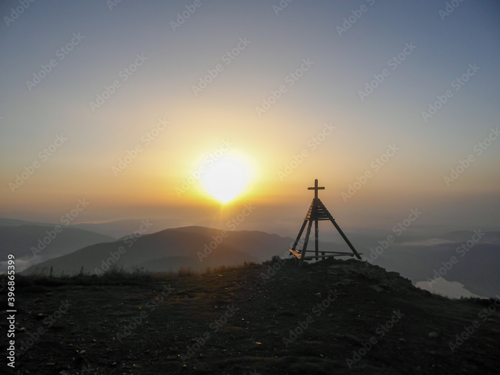 A panoramic view on a sunrise with small tower with a cross on the top on the peak of Gerlitzen, Austrian Alps. The skyline is exploding with orange. The valley is shrouded in fog. Daybreak