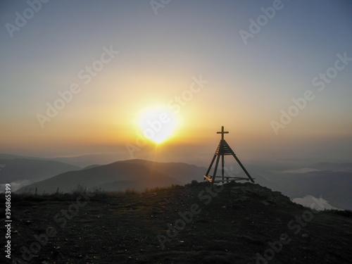 A panoramic view on a sunrise with small tower with a cross on the top on the peak of Gerlitzen, Austrian Alps. The skyline is exploding with orange. The valley is shrouded in fog. Daybreak