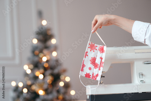 A mid shot of a Christmas designed handmade face mask held by a girl's hand. Christmas tree and sewing machine on the background photo