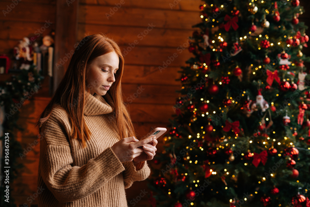 Side view of serious redhead young woman typing messages on mobile phone near Christmas tree. Middle shot portrait of female chatting online on cellphone at cozy living room with festive interior