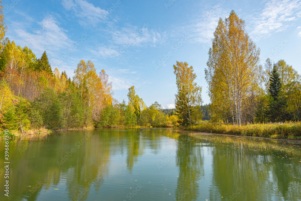 Autumn forest trees are reflected in the river water of the panoramic landscape. Blue sky with clouds. Autumn forest trees are reflected in the river water of the panoramic landscape. Blue sky with clouds.