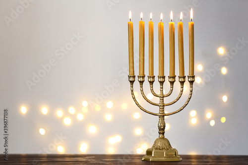 Golden menorah with burning candles on table against light grey background and blurred festive lights, space for text