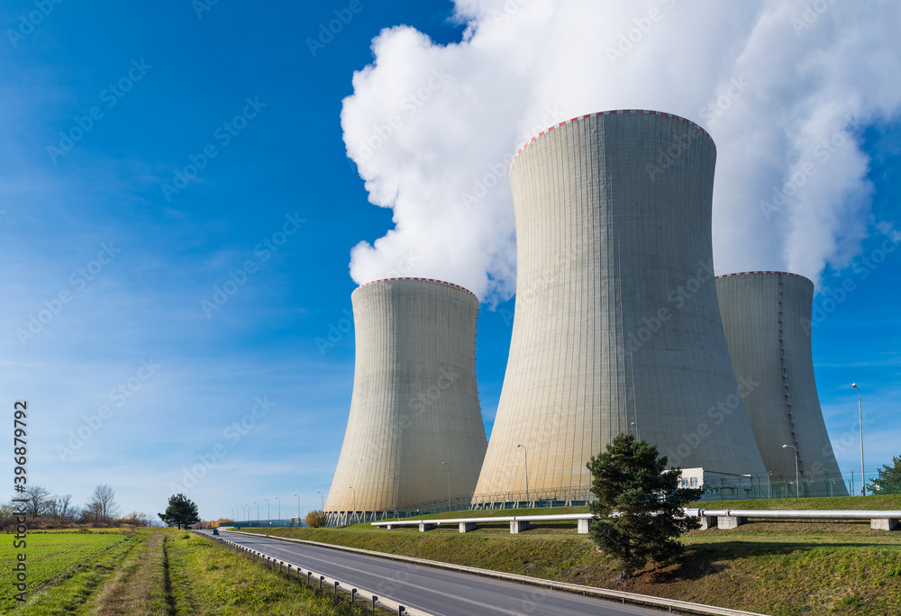 Nuclear power plant cooling towers with water vapor plumes on blue sky background. Closeup of modern generating station in landscape with a road and steam piping on green grass. Eco. Temelin, Czechia.