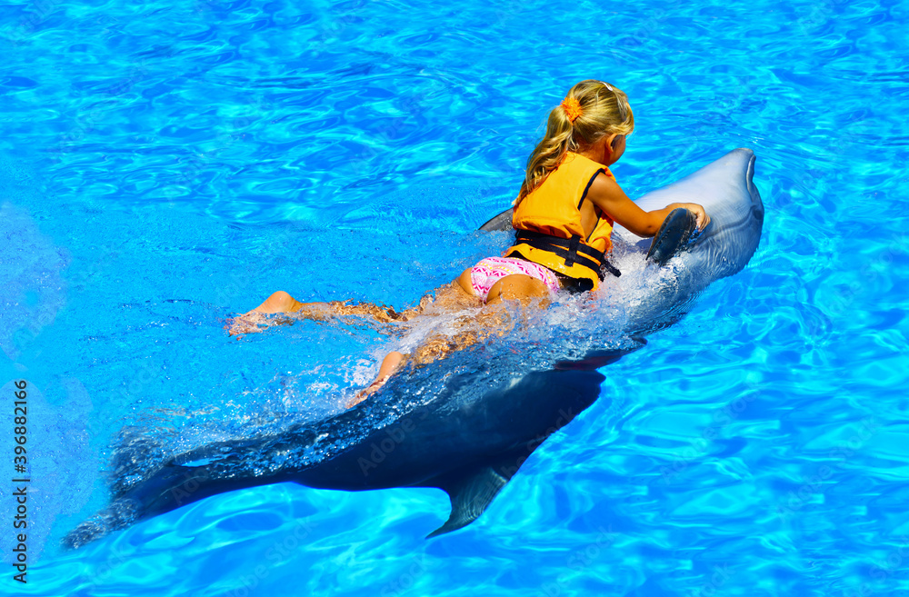 A little girl riding a dolphin rides in the dolphinarium with blue and transparent water. Joy. Pleasure. Water splashes and bubbles. The child plays with a cute sea whale, holding tightly to the fins.