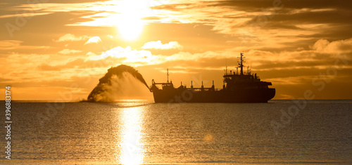 Vessel engaged in dredging at sunset time. Hopper dredger working at sea. Ship excavating material from a water environment. Beautiful sunset. photo