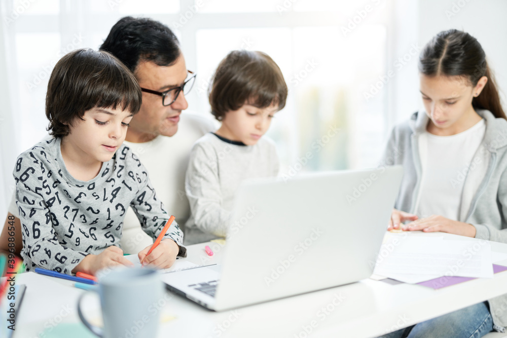 Caring latin father spending time with his children. Dad using laptop, working from home and watching kids drawing while sitting at the table with him Caring latin father spending time with his children. Dad using laptop, working from home and watching kids drawing while sitting at the table with him