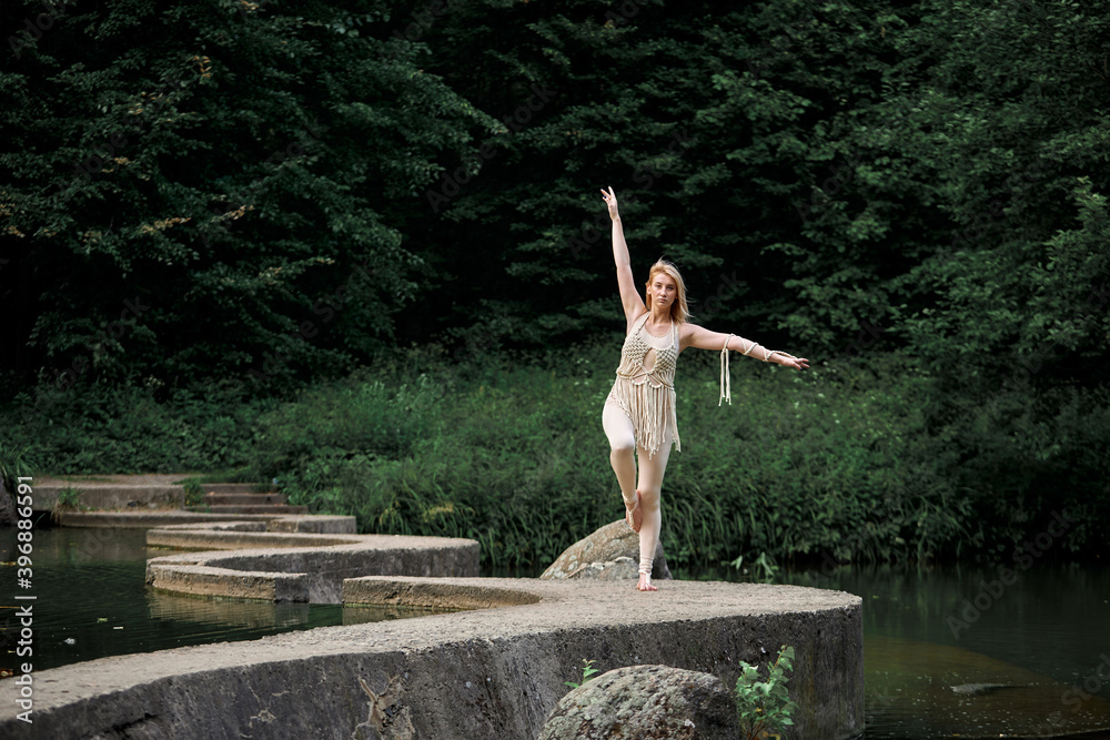 A young blonde woman stands barefoot on one leg on a winding concrete bridge. Clothing of their eco-materials, macrame. The concept of natural products.