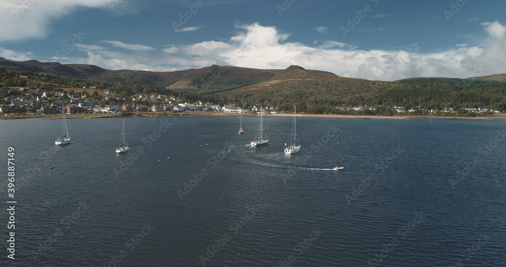 Yacht racing at ocean bay coast aerial. Passengers on sailboat at open sea at summer cloudy day. Cinematic scenery of luxury cruise on sail boat. Brodick harbor at mountain island of Arran, Scotland