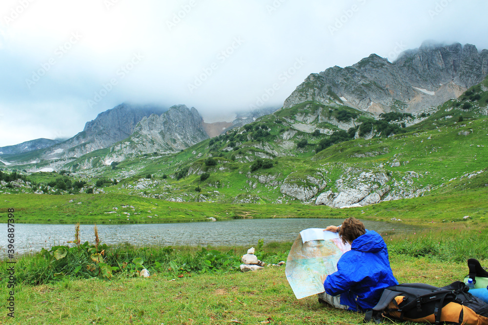 Person with a backpack in the mountains. A hiker in a blue raincoat looks at a geographical paper map. Woman tourist near the lake