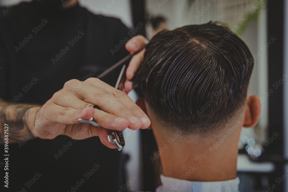 detail of a young man with tattooed arms cuts a man's hair in a barber shop