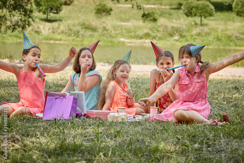 Four girls in holiday hats celebrate a friend's birthday in nature in the summer. Gifts in packages. Picnic with sweets.