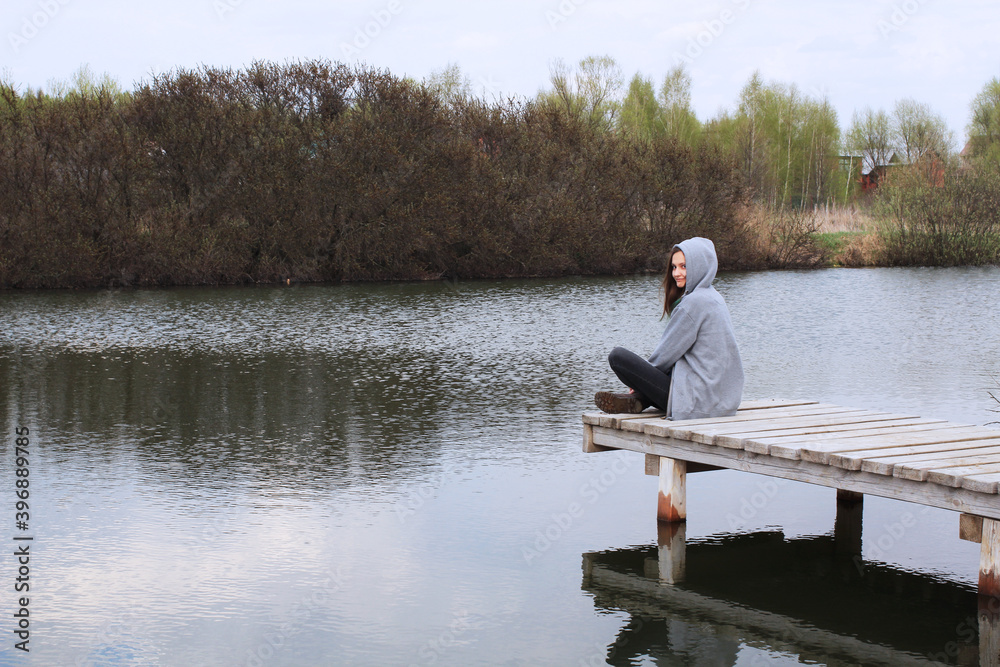 Countryside. Young woman resting sitting on the pier near the lake. Person in a hoodie with a hood