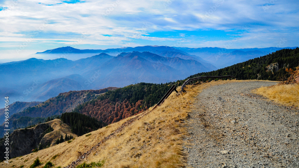A gravel road winding along Cozia Peak. Olt valley lies bellow and Capatanii Mountains can be seen along the horizon. Hazy clouds are covering the mountain peaks.
