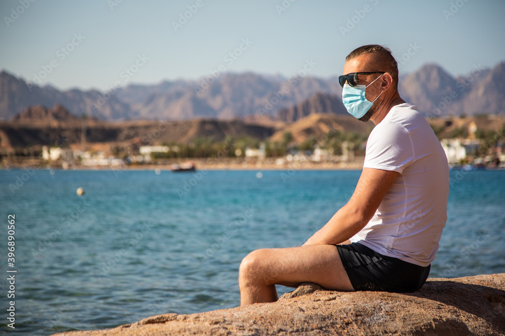 tourist in a protective mask on vacation. man in a medical mask sits on the seashore