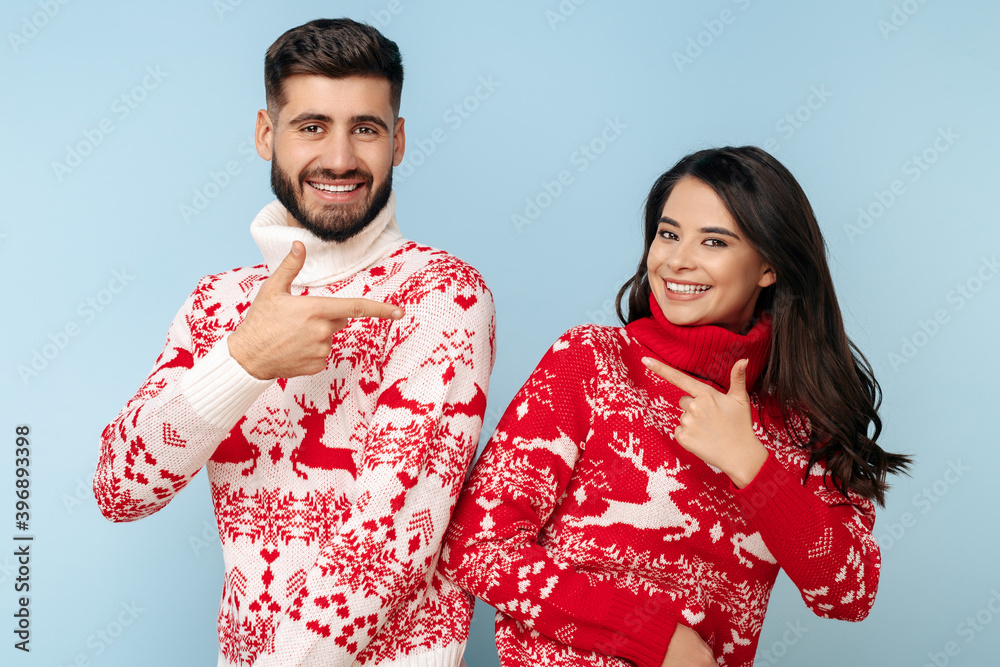 Joyful couple dressed in Christmas sweaters pointing fingers smiling over blue background.
