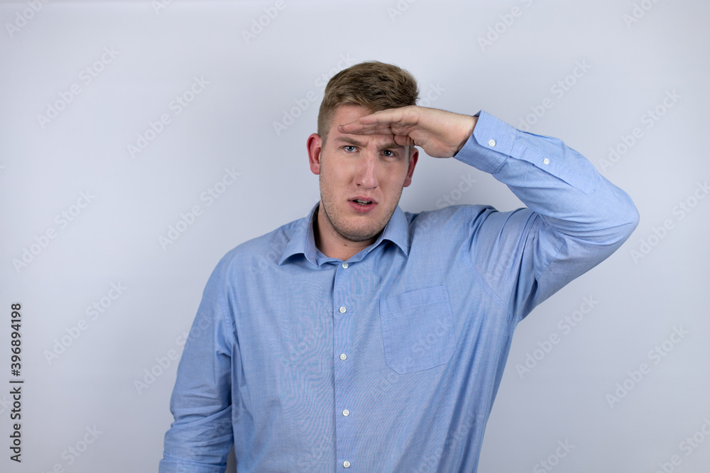 Business young man wearing a casual shirt over white background very happy and smiling looking far away with hand over head. searching concept. Business young man wearing a casual shirt over white background very happy and smiling looking far away with hand over head. searching concept.