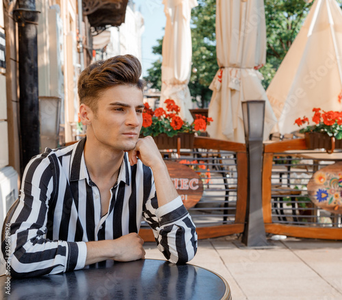 Guy model near the cafe. A young man in stylish clothes is sitting at a table. Cute guy on the summer terrace with flowers.