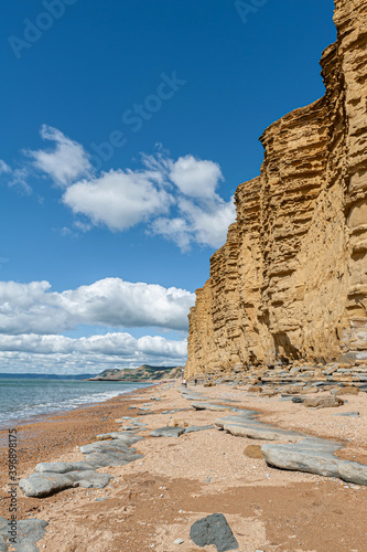 People walking on golden beach underneath towering cliffs on sunny summer day. Jurassic coastline of West Bay in Dorset. UK