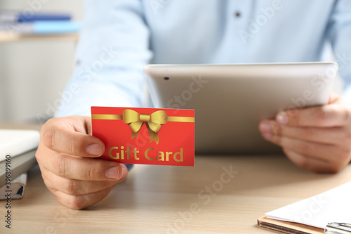 Man with gift card and tablet at table indoors, closeup