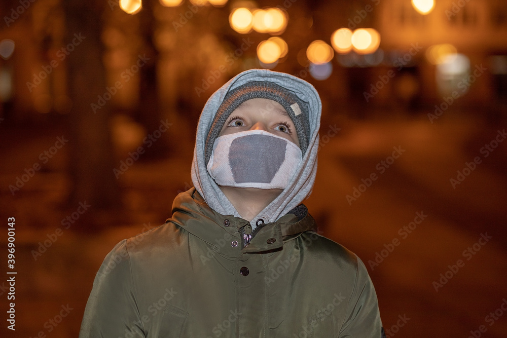 Close up portrait of Caucasian teenager boy in mask and warm casual clothes standing on street looking up. Little male teenager in city night lights. Close up portrait of Caucasian teenager boy in mask and warm casual clothes standing on street looking up. Little male teenager in city night lights.