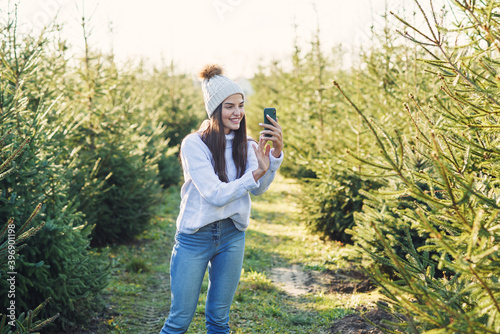 Joyful pretty young woman in warm hat photography beautiful christmas trees during walking among forestry plantings. © gorynvd