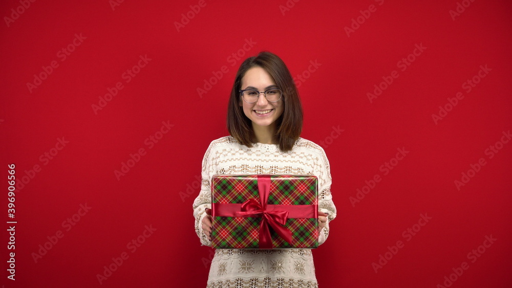 A young woman with glasses shakes a Christmas present in her hands and rejoices. Shooting in the studio on a red background. A young woman with glasses shakes a Christmas present in her hands and rejoices. Shooting in the studio on a red background.