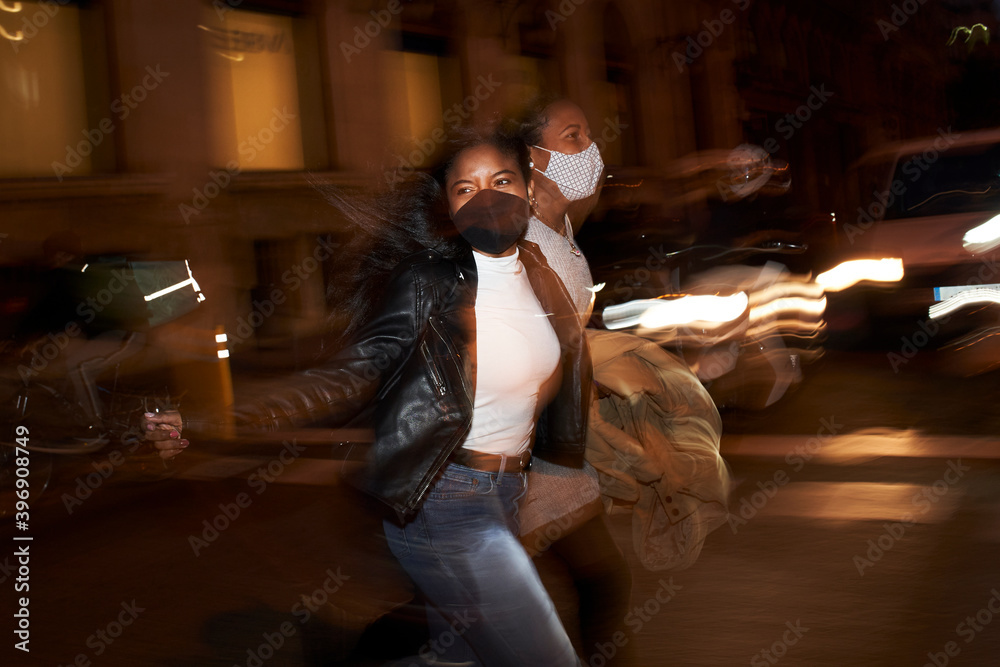 Flash portrait of two African American women with face masks running down the street and laughing at night in a busy city. City lights and motion blur