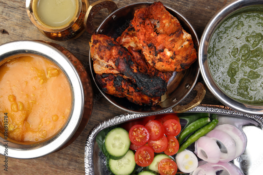 Spicy red Chicken grilled tikka tandoori nan bread green spinach curry yogurt sauce dal tomato cucumber onion salad set in metal stainless steel brass copper pot on rustic vintage wood background