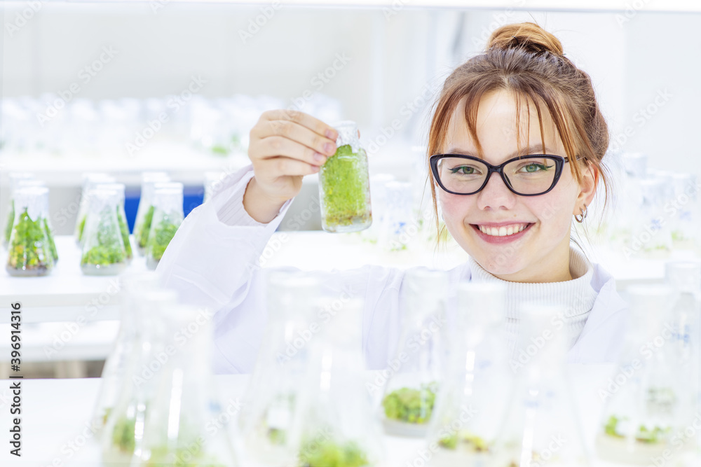 A girl laboratory assistant in a biological laboratory holds a flask with plants grown in a nutrient in her hands with a smile looks at the camera. Biological research at the university A girl laboratory assistant in a biological laboratory holds a flask with plants grown in a nutrient in her hands with a smile looks at the camera. Biological research at the university