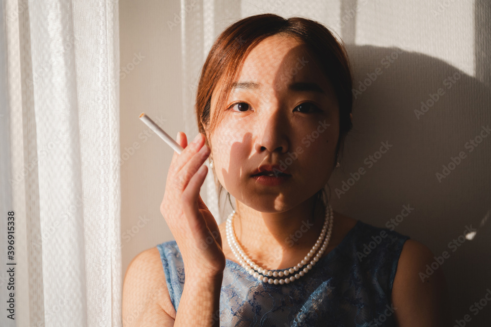 部屋の窓際でタバコを持つ青いドレスを着た若い女性 部屋の窓際でタバコを持つ青いドレスを着た若い女性