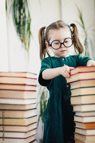 Obraz na plátne Little cute preschool age girl in round shaped glasses between books pile