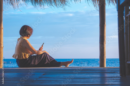 Side view of Asian woman is relaxing and using smartphone in cottage on seaside against seascape view background during her weekend