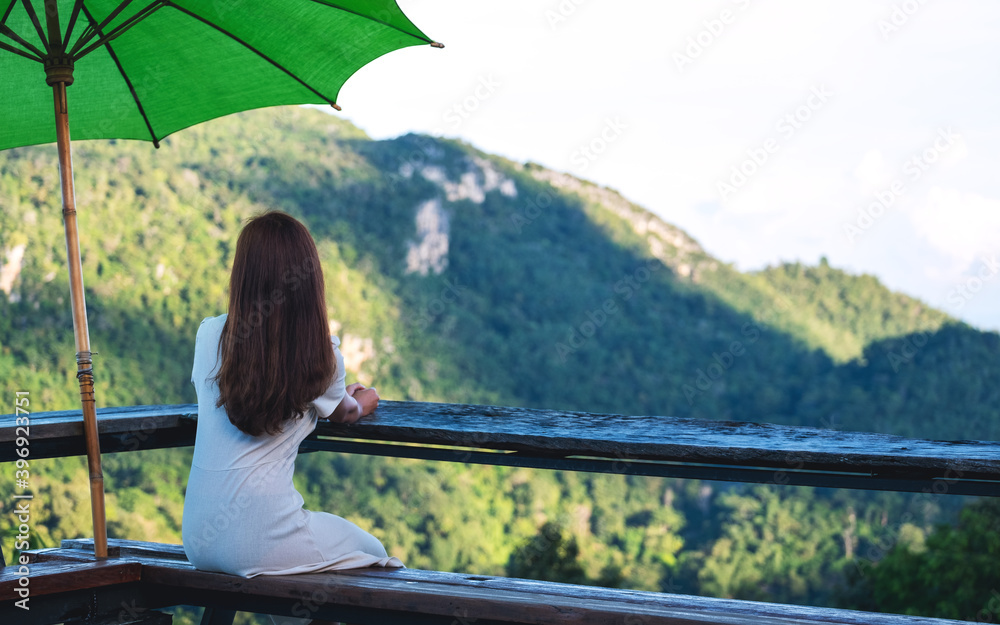 Rear view image of a female traveler sitting on wooden balcony and looking at a beautiful mountain and nature view