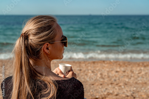 A beautiful blonde with long hair gathered in a ponytail sits on the beach drinking coffee and enjoying the views. photo