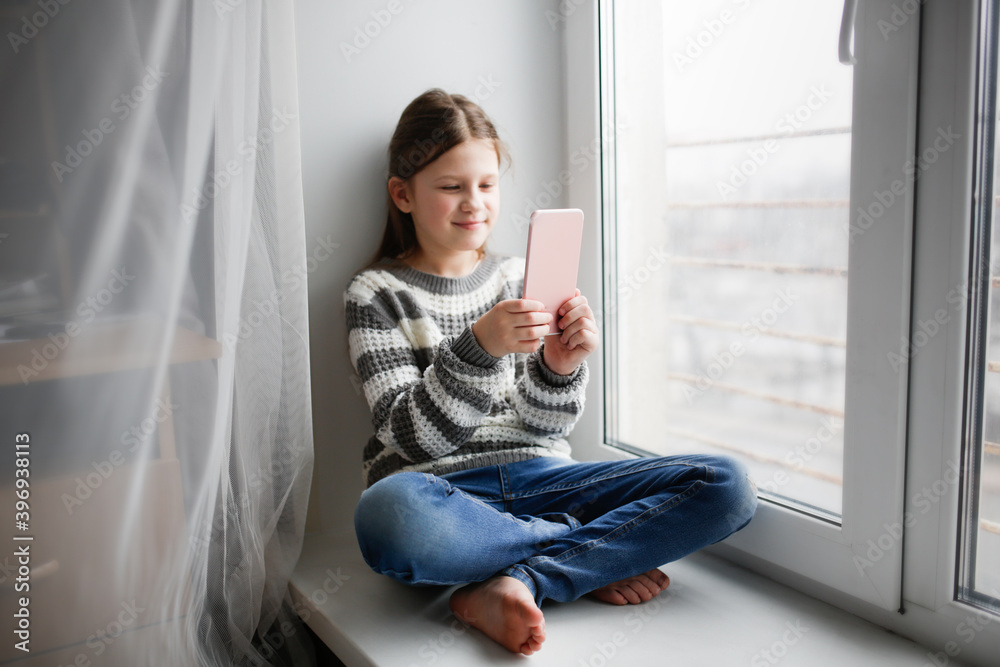 Cute caucasian child girl 8 9 years on the windowsill makes a selfie with filter, talks on video communication, plays with a smart phone. Kids and gadgets concept Cute caucasian child girl 8 9 years on the windowsill makes a selfie with filter, talks on video communication, plays with a smart phone. Kids and gadgets concept